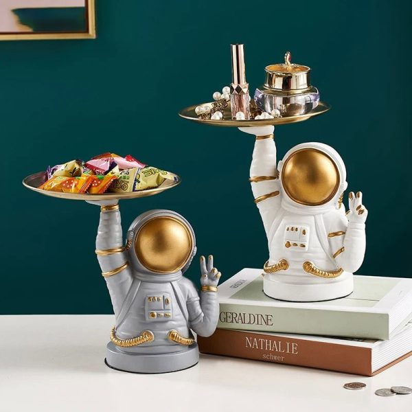 Figura de astronauta em cerâmica branca e dourada, usada como decoração, segurando uma bandeja com objetos de decoração e doces, estilo moderno e original.