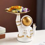 Figura de astronauta em cerâmica branca com capacete dourado, segurando uma bandeja cheia de doces coloridos, decoração original para um espaço temático ou uma coleção de objetos científicos.