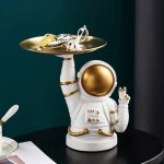 Figura de astronauta em cerâmica branca e dourada, segurando uma bandeja com acessórios científicos, decoração de estilo cósmico para coleção ou decoração.