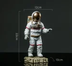 Figura de astronauta em miniatura com capacete dourado, medindo 22 cm de altura, 12 cm de largura e 10 cm de profundidade, colocada sobre uma base rochosa, ideal para decoração científica.