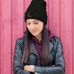 Jovem mulher usando um gorro preto com padrão lunar e estrelas, vestida com uma jaqueta de couro preta e um moletom com capuz roxo, posando em frente a uma parede de madeira rosa.