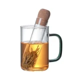 Caneca de vidro com infusor de chá, cheia de água quente infundindo folhas de chá, com tampa de cortiça colocada no topo.