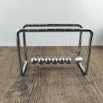 Pêndulo de Newton em metal brilhante com seis esferas suspensas, ilustrando a conservação de energia e a física newtoniana.