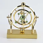 Modelo atômico decorativo em metal dourado com esferas verdes e douradas, representando um átomo estilizado.