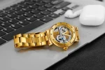 Relógio mecânico dourado com movimento visível, colocado sobre um laptop preto com fones de ouvido brancos ao lado.