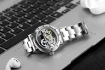 Relógio mecânico com movimento visível, pulseira em aço inoxidável, colocado sobre um teclado de computador.