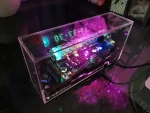 Painel de exibição eletrônico LED em caixa transparente, exibindo números luminosos em azul e verde, com componentes eletrônicos visíveis no interior.