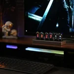 Dispositivo de tubos de vácuo luminosos com números e símbolos, colocado sobre uma mesa com um ecrã de computador ao fundo, em ambiente escuro.
