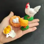 Poule en plastique blanc avec crête rouge, œufs blancs dans un nid orange, petite poule jaune sur un œuf cassé, figurines décoratives pour thème scientifique ou éducatif.