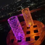 Dois vasos de cristal luminosos, um roxo e outro laranja, colocados sobre uma mesa de madeira com vista para a cidade iluminada à noite.
