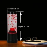 Vaso de bolhas de sabão com luz LED vermelha, altura 25,5 cm e diâmetro 9 cm, colocado sobre uma mesa de madeira com óculos e um caderno ao lado, fundo preto.