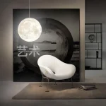Lâmpada de parede em forma de lua suspensa acima de uma poltrona moderna branca em um interior contemporâneo, com prateleira ao fundo.