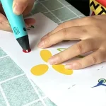 Pinça de desenho para crianças usando um marcador luminoso para criar formas coloridas em uma folha de papel. Ideal para estimular a criatividade e o aprendizado lúdico.