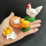 Galinha de plástico branco com crista vermelha, ovos brancos em um ninho laranja, pequena galinha amarela sobre um ovo quebrado, figuras decorativas para tema científico ou educativo.