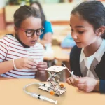 Duas jovens exploram um robô educativo científico durante uma atividade em sala de aula, ilustrando o aprendizado lúdico de tecnologia e robótica para crianças.