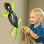 Criança pequena sorridente segurando um controle remoto, interagindo com um lagarto robô luminoso fixado na parede.
