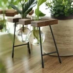 Jardineira suspensa com plantas verdes em um vaso de vidro, suporte de madeira e metal, decoração interior natural e moderna.