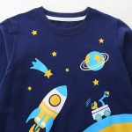 Roupa para criança com padrão espacial, incluindo um foguete, estrelas, um planeta e um telescópio, perfeita para amantes de astronomia e exploração espacial.