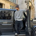 Homem em traje casual posando em frente a um carro preto numa rua urbana.